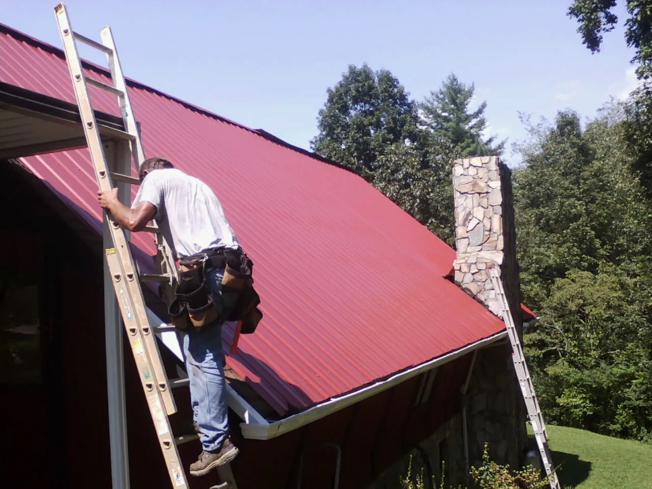 Roofer on a ladder beside a red metal roof and stone chimney — Soffit & Fascia Repair by Roofing Brothers in Almond